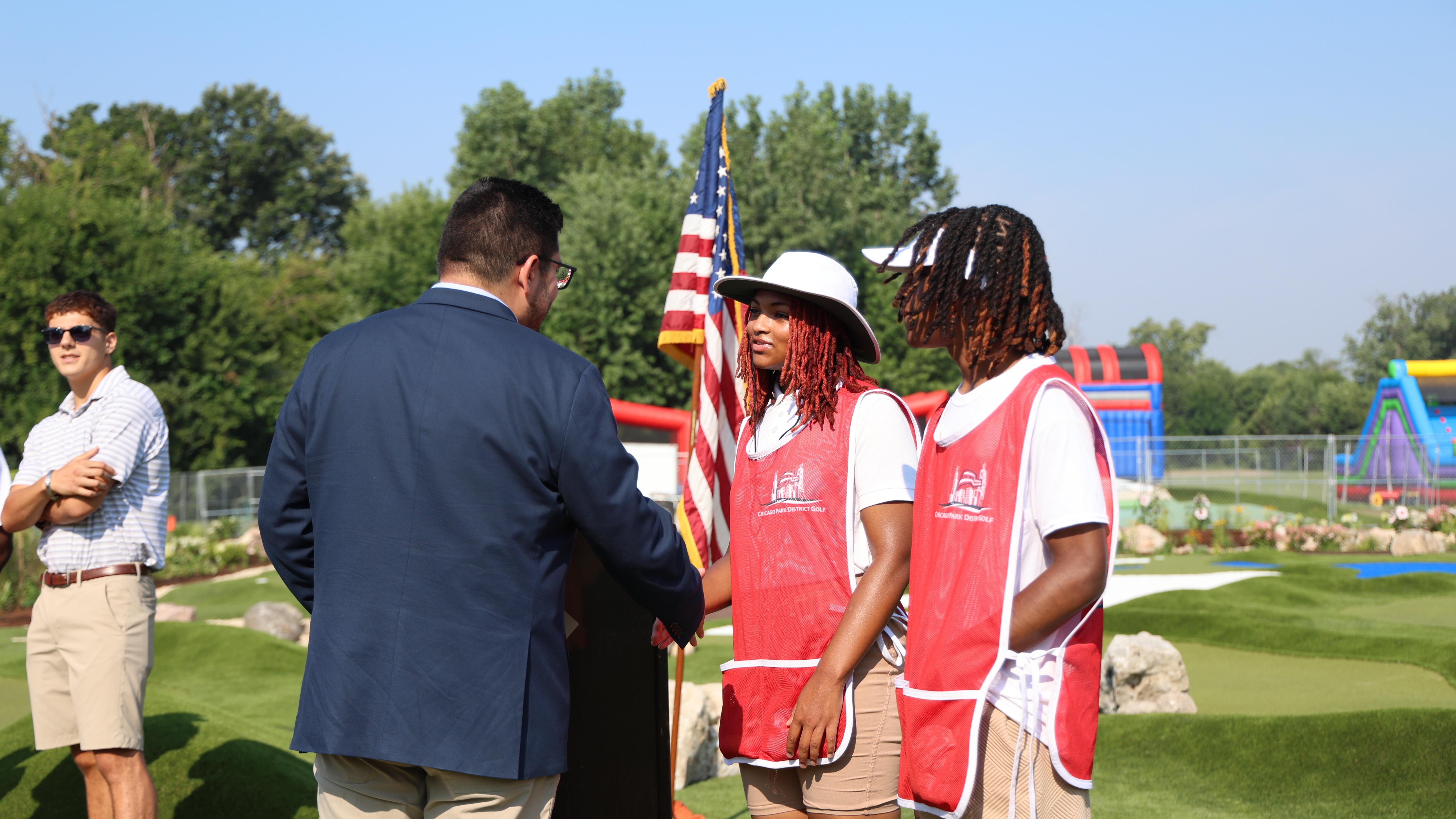 People in red golf vests talk with a man in a suit near a US flag at a mini golf course.